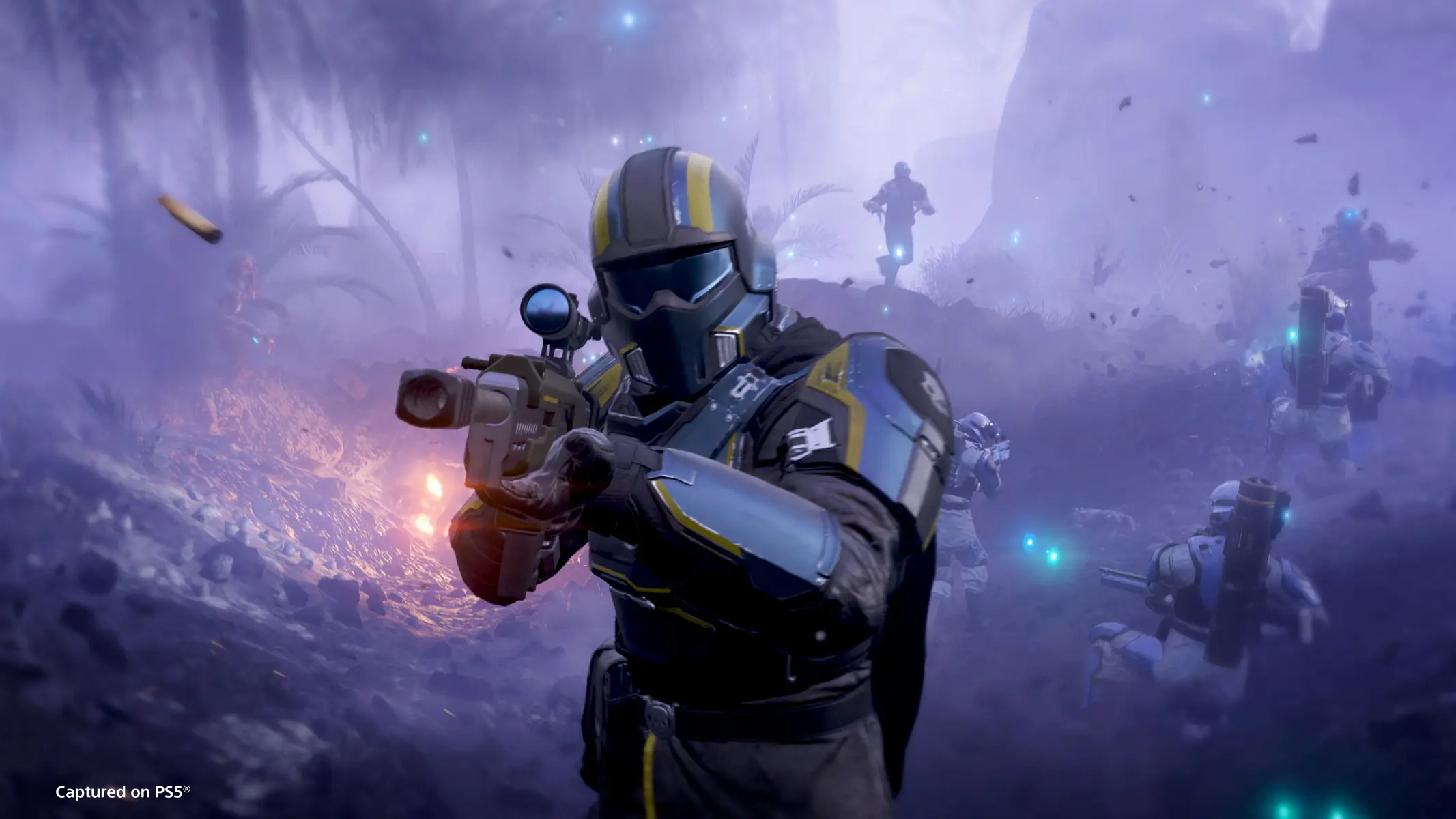 A Helldiver in blue and yellow armor aiming a rifle during a chaotic battle in Helldivers 2.